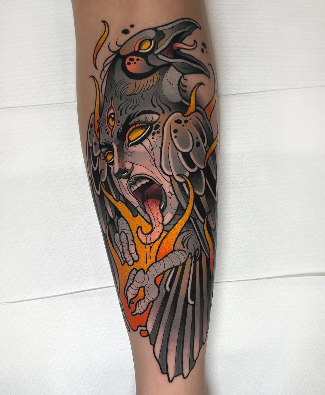 Colorful leg tattoo featuring a woman's face with bird and animal elements, flames, and golden yellow eyes on the calf.