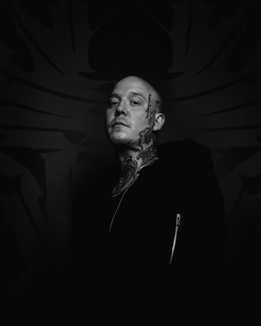 Bald man with extensive neck and head tattoos wearing a black leather jacket, photographed in black and white against a dark background.