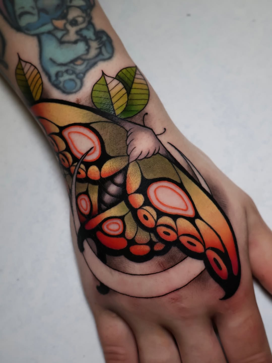Colorful butterfly tattoo on the hand featuring red and orange wings with green leaves, black outlines, and detailed wing patterns.