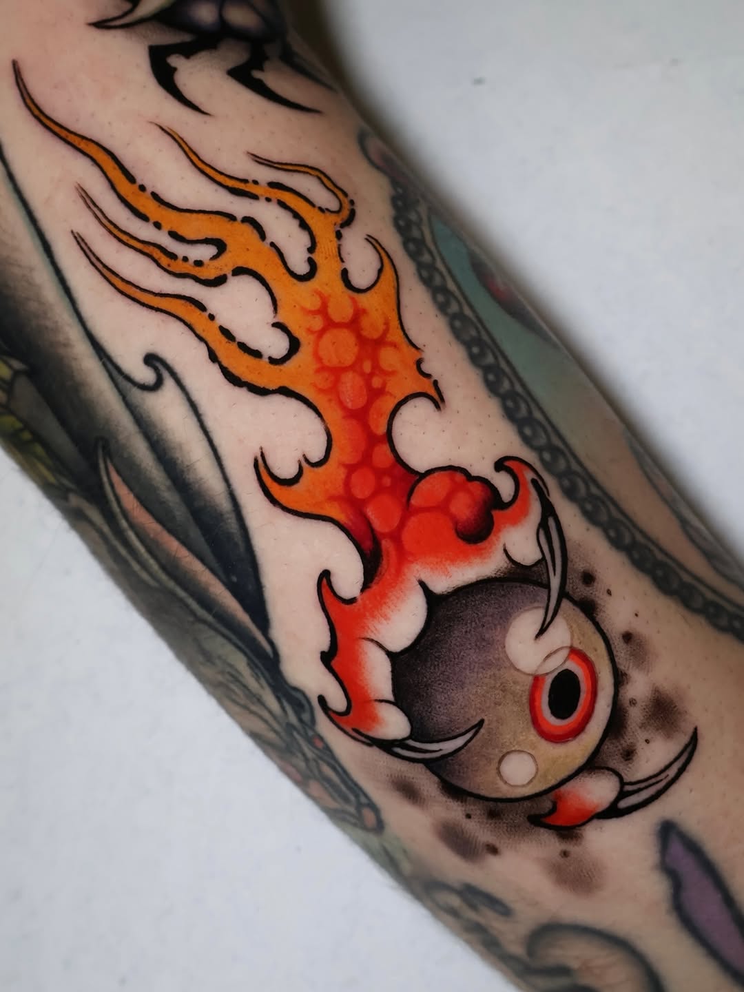 Colorful koi fish tattoo with orange, red, and black details on an arm, surrounded by decorative flame-like elements and dotted patterns.