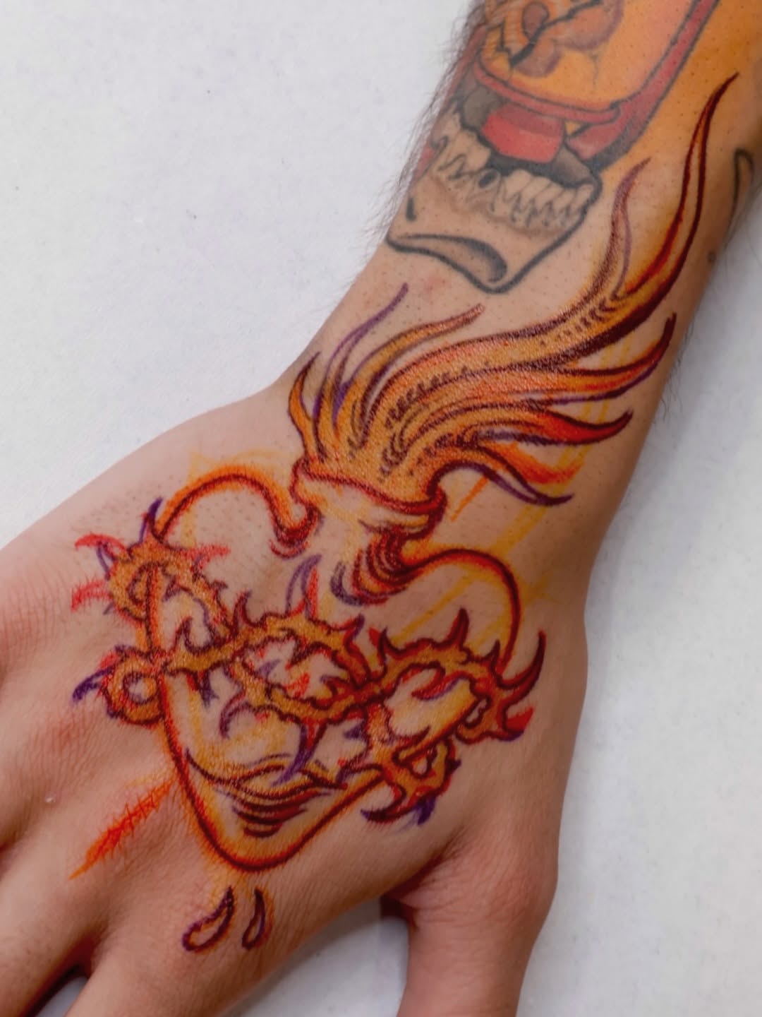 Colorful phoenix tattoo on hand and forearm featuring red, orange, yellow, and purple flames with a skull design on the upper arm.
