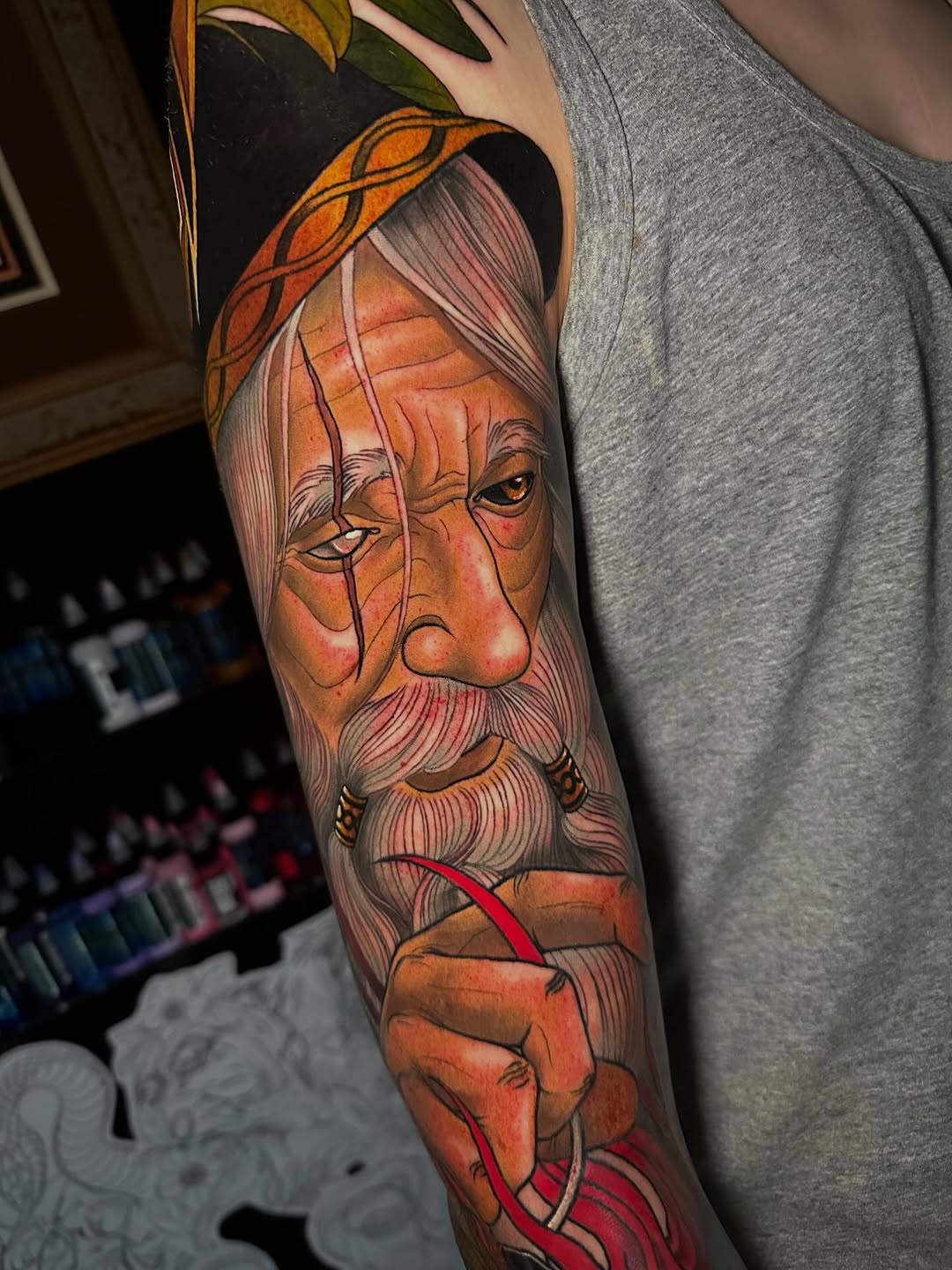 Colorful arm sleeve tattoo depicting an elderly bearded man with orange skin tones, headwrap, and ornamental details on the upper arm.