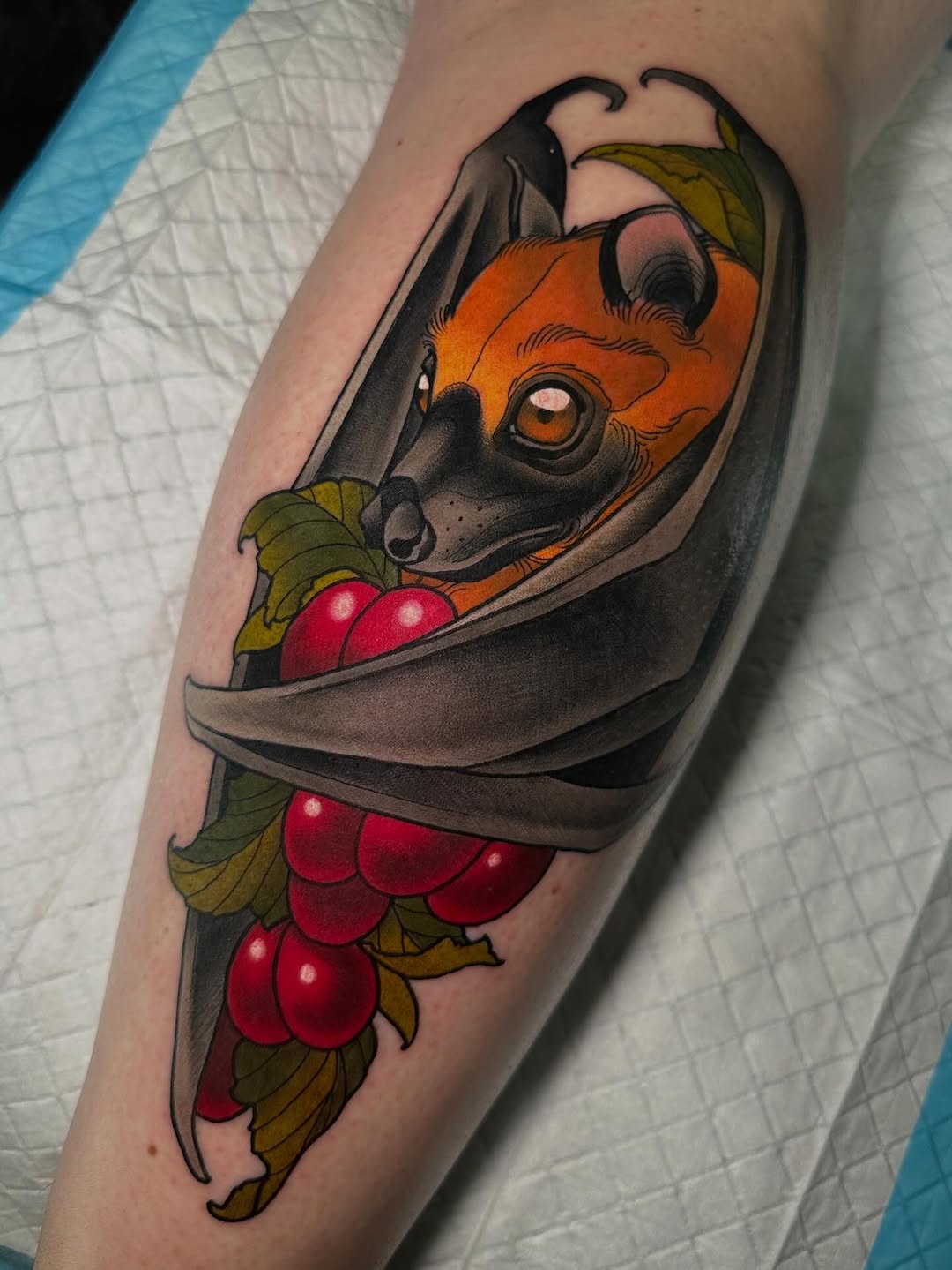 Colorful tattoo on an arm featuring an orange-faced devil with horns wrapped in gray fabric, surrounded by red berries and green leaves.