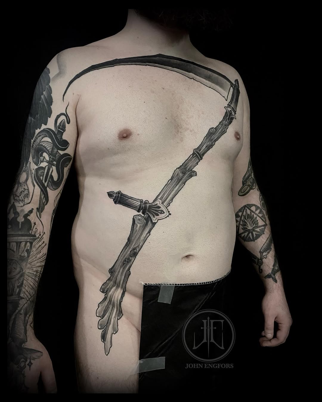 Shirtless male torso displaying a large detailed sword tattoo across the chest and shoulder, with additional designs on both arms.