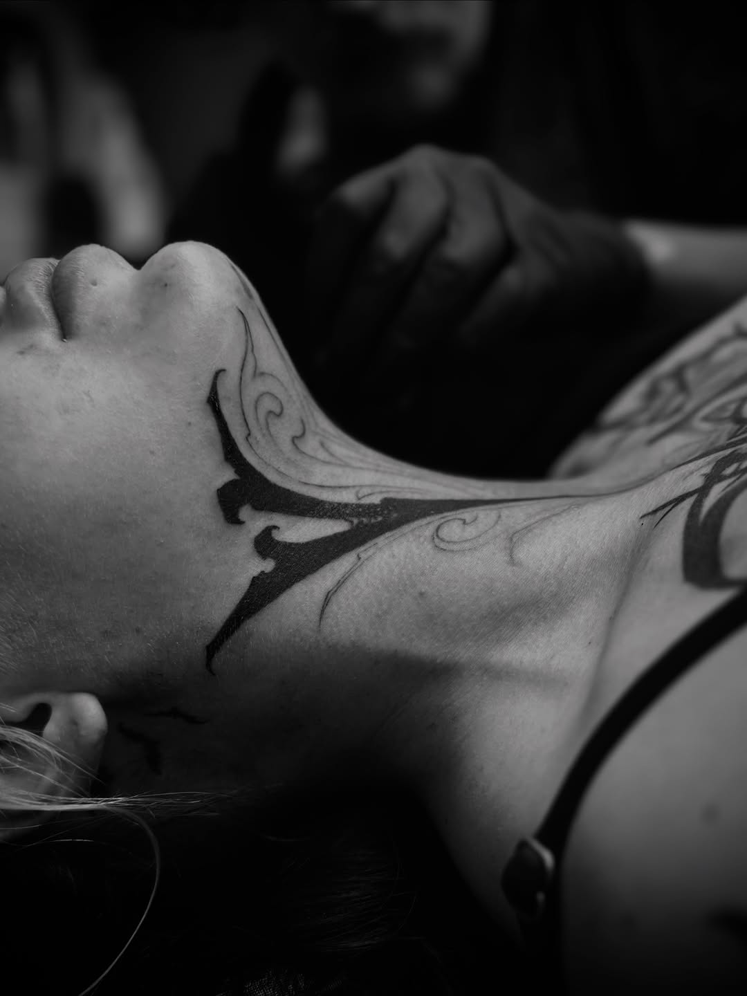 Black and grey ornamental swirl tattoo on the neck and shoulder of a person with dark hair, photographed in profile.