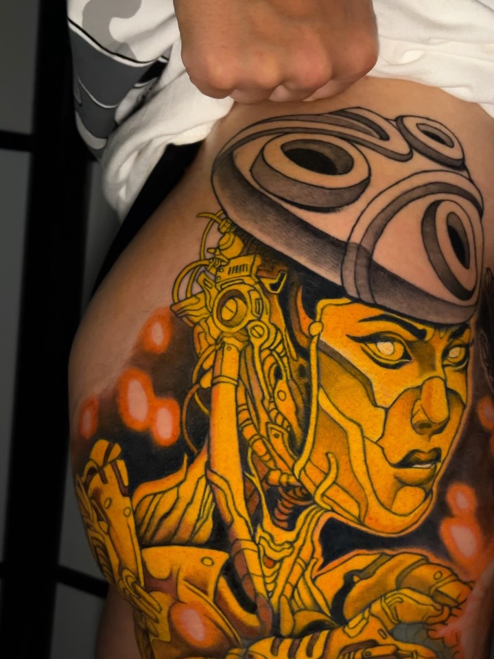 Colorful shoulder tattoo featuring a golden-faced figure with mechanical headpiece and orange circular elements on dark background.