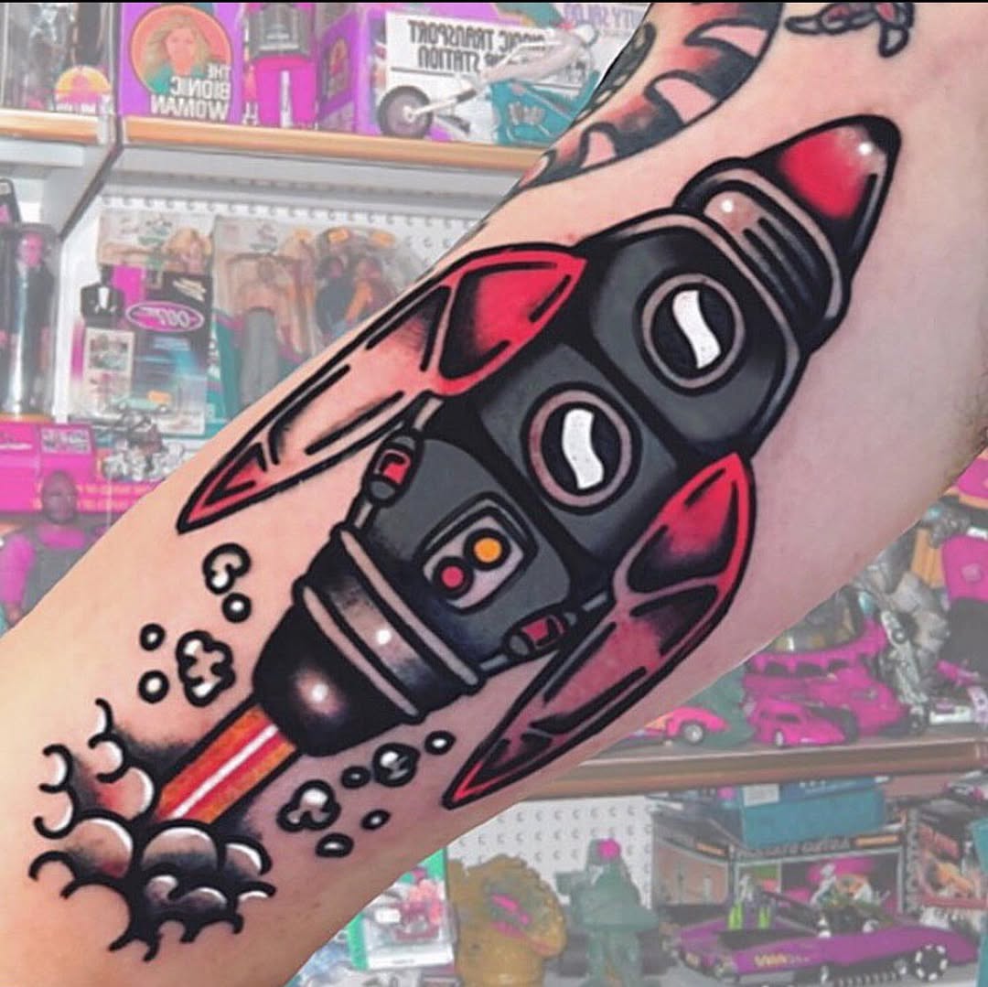 Red and black rocket ship tattoo with exhaust clouds on an arm, displayed against a colorful toy store backdrop.