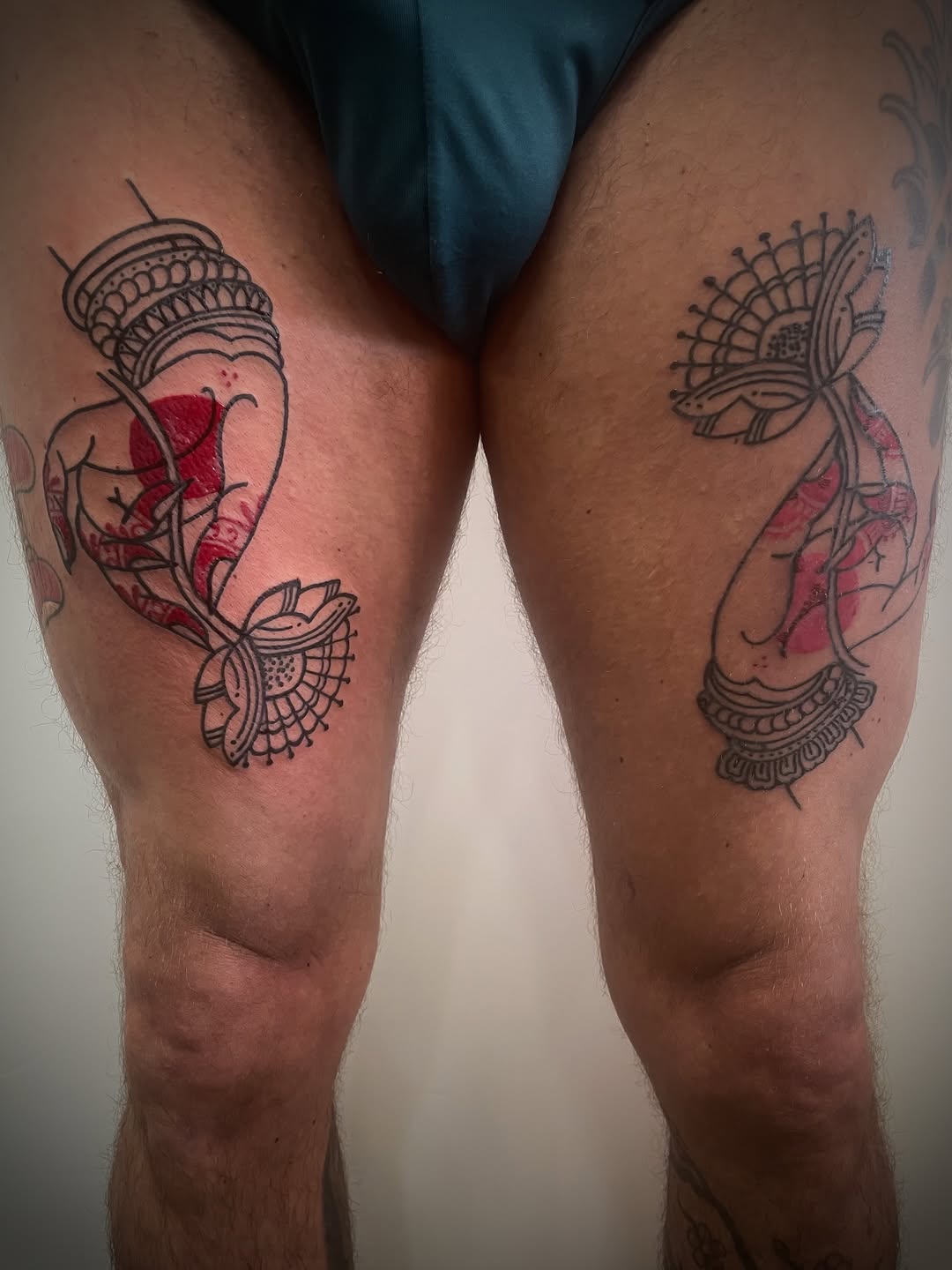 Matching leg tattoos featuring ornate fan designs with red watercolor accents and decorative linework on both thighs.