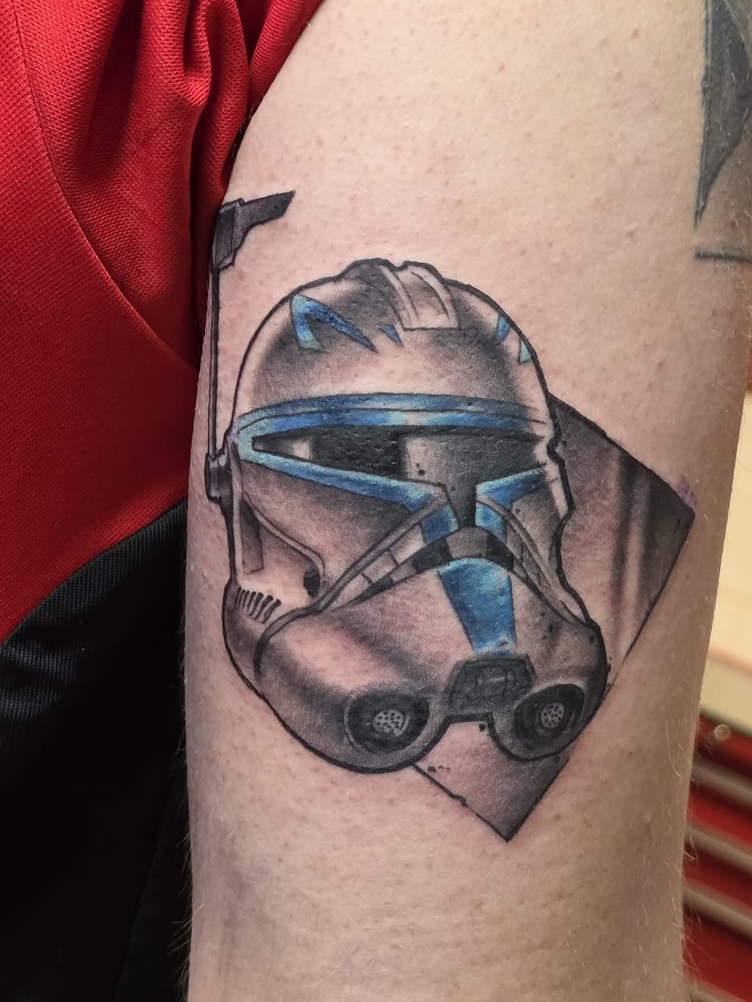 Close-up of a stormtrooper helmet tattoo on an arm with grey shading, blue accents, and black details against skin.