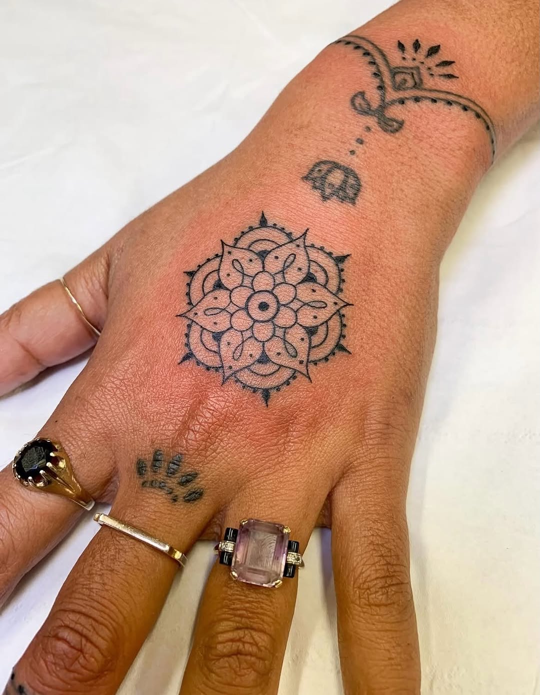 Hand tattoo featuring a mandala-inspired flower design on the back of the hand, with additional small symbols on the forearm and wrist, done in black ink.