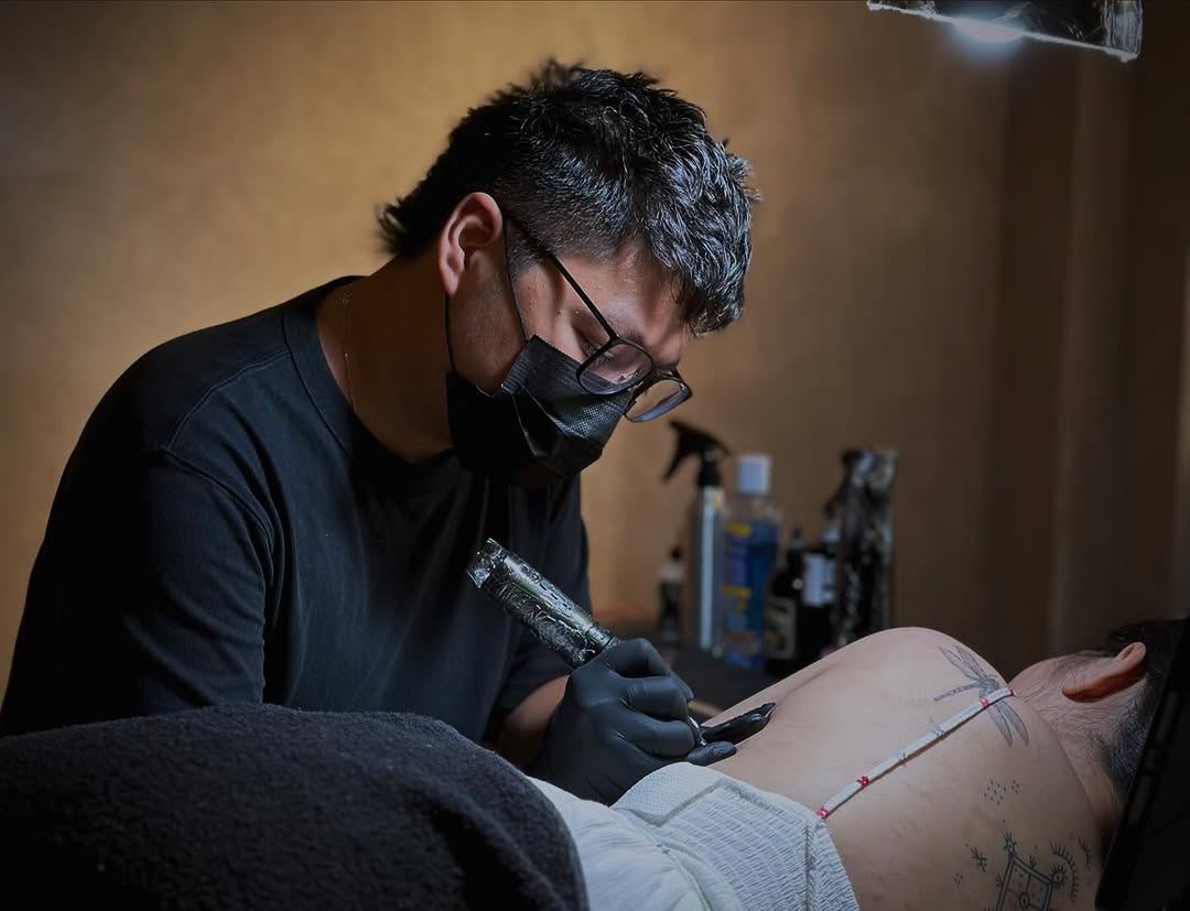 A tattoo artist wearing glasses and a black mask applies ink to a client's arm during a session in a studio setting.