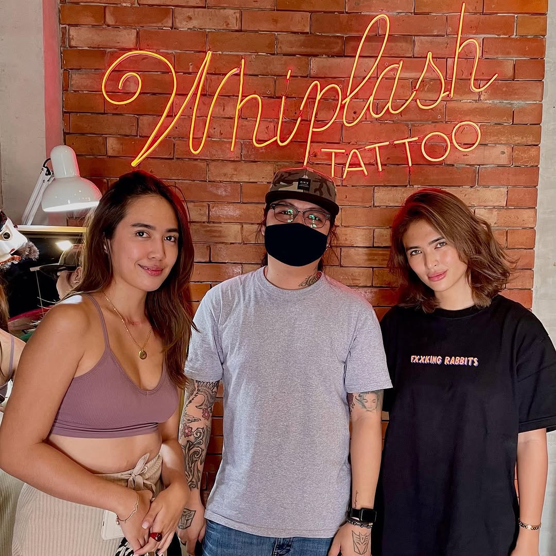 Three people pose in front of a neon sign at a tattoo studio, displaying various visible tattoos on their arms and bodies in different colors and designs.