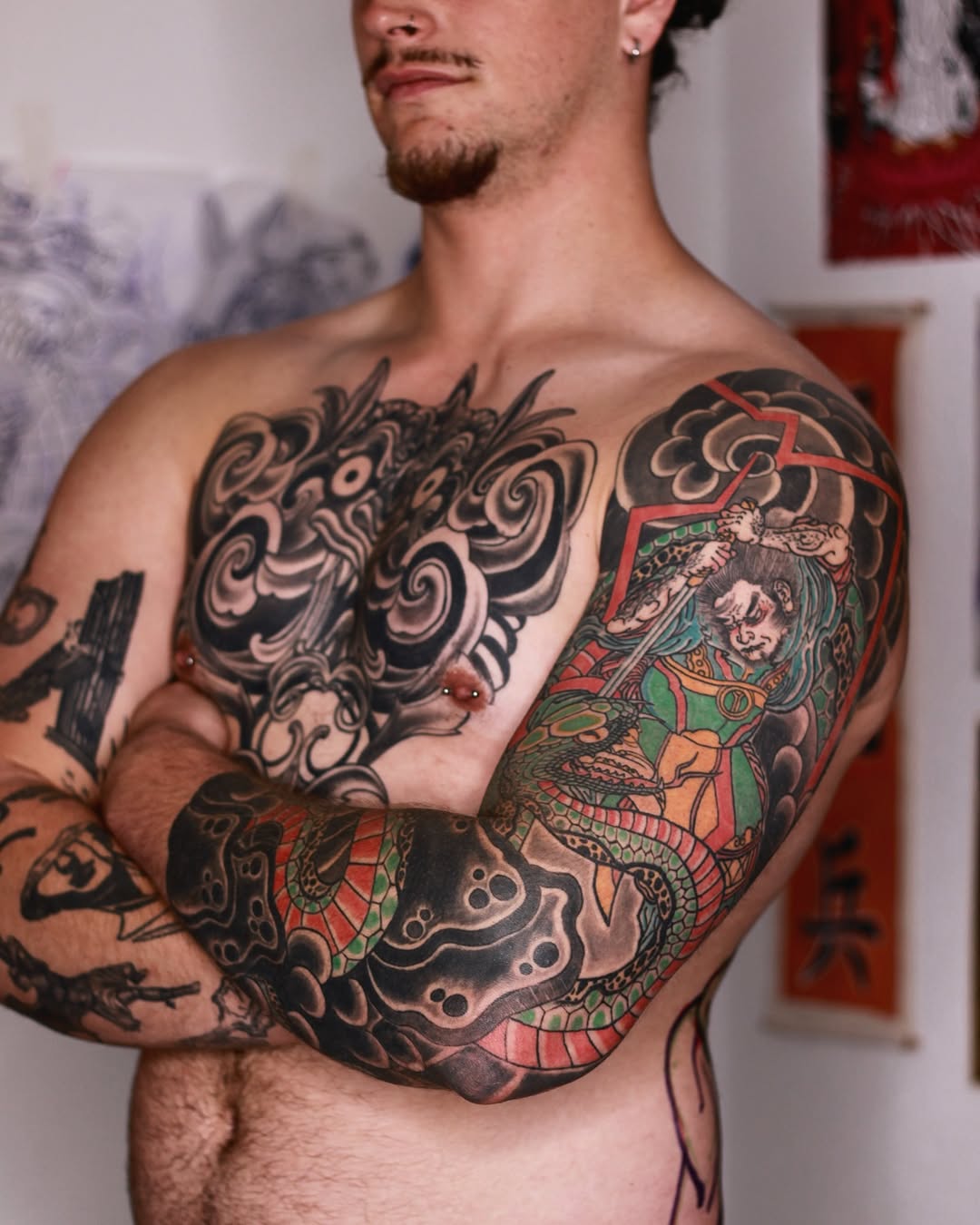 Shirtless man displaying large colorful chest and sleeve tattoos featuring ornate designs, dragons, and figures in red, green, black, and skin tones.