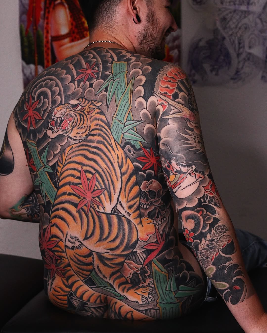 Back tattoo featuring a large orange tiger with black stripes, surrounded by clouds, red flowers, green bamboo, and skulls in orange, red, and neutral tones.