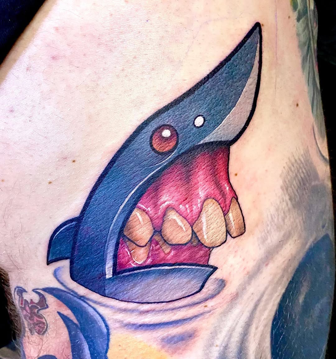 Colorful tattoo on upper arm featuring a blue shark with red mouth, tan teeth, and white eye highlights against pale skin.