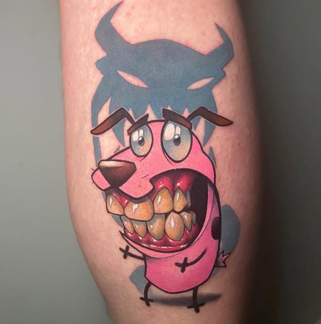 Colorful tattoo on upper leg featuring a pink creature with large eyes, prominent teeth, brown horns, and a dark blue demonic face looming above it.