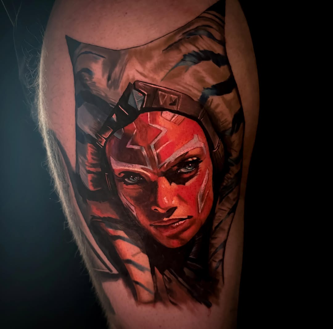 Colorful shoulder tattoo featuring a red-faced figure with geometric markings and a dark silhouetted face in the background.