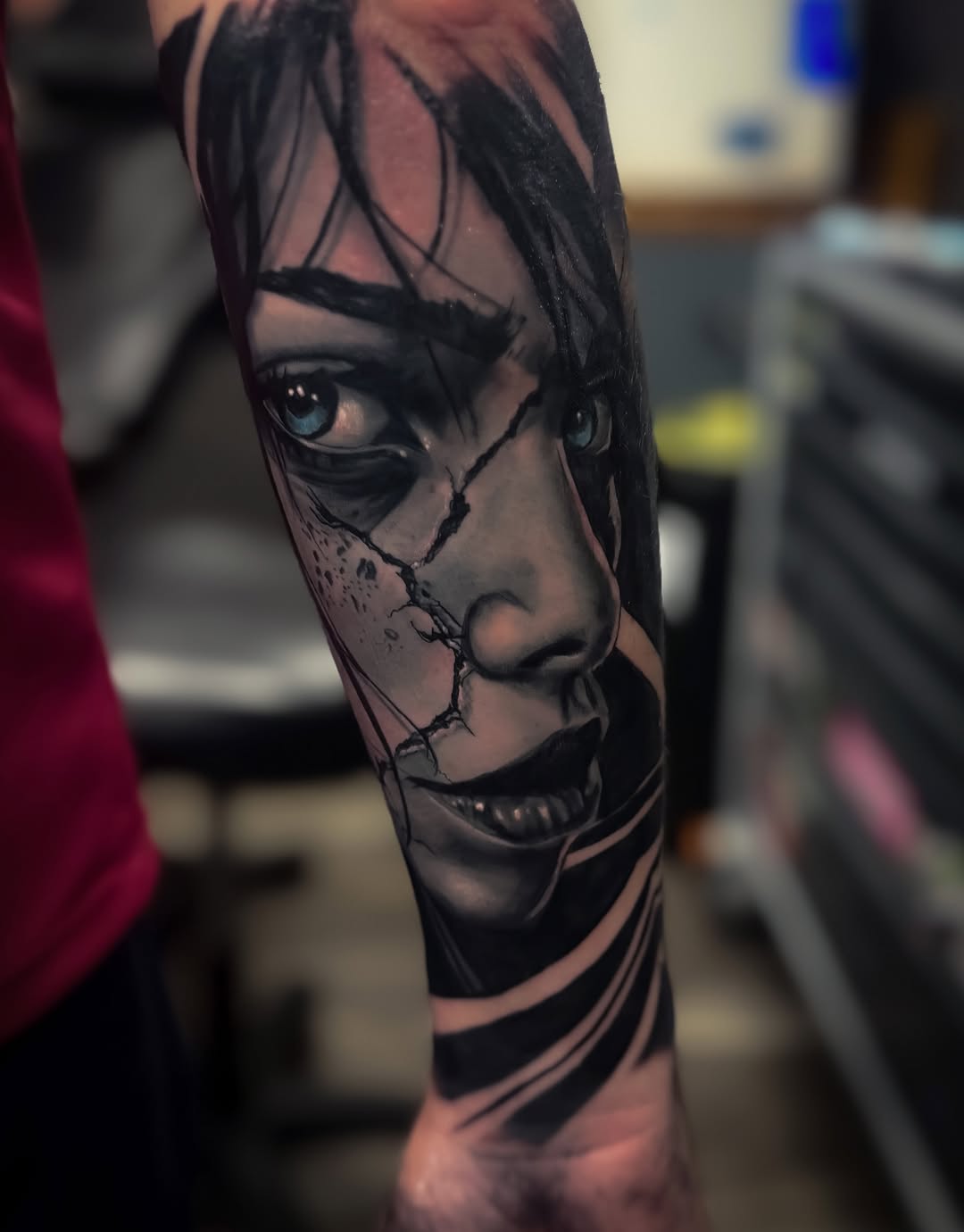 Detailed portrait tattoo of a face with striking eyes and stitched details on the forearm, rendered in black ink with shading on tan skin.