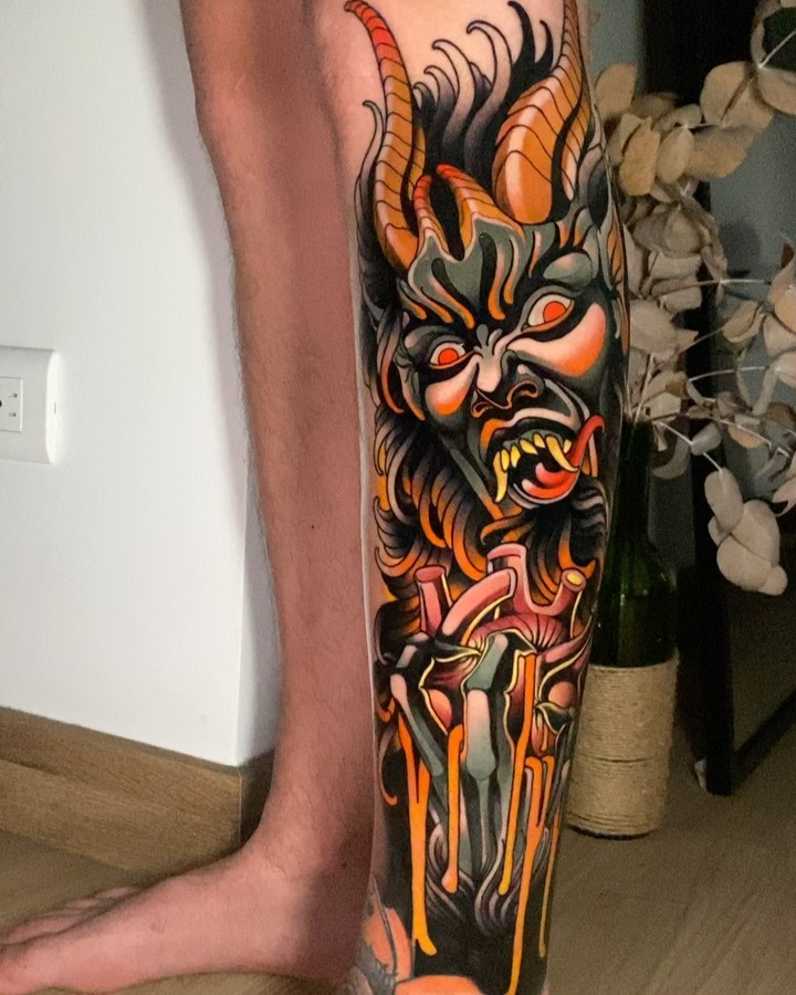Colorful demon head tattoo with orange and black horns, glowing red eyes, and a sword piercing through the neck on the calf.
