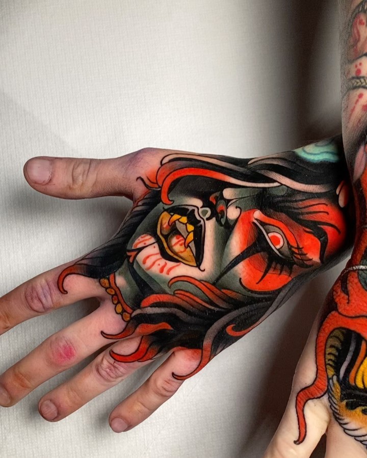 Colorful demon or oni mask tattoo on hand and fingers featuring red, black, yellow, and orange tones with flowing hair details.