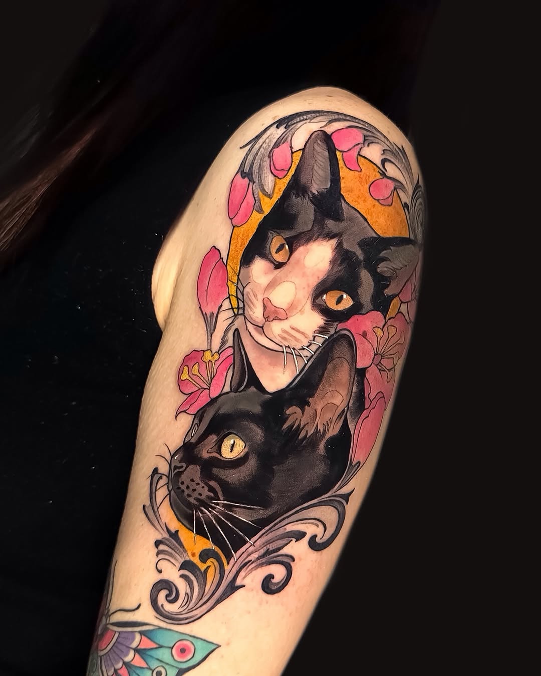 Colorful tattoo on upper arm featuring two black cats with golden eyes surrounded by pink and orange flowers and decorative scrollwork.