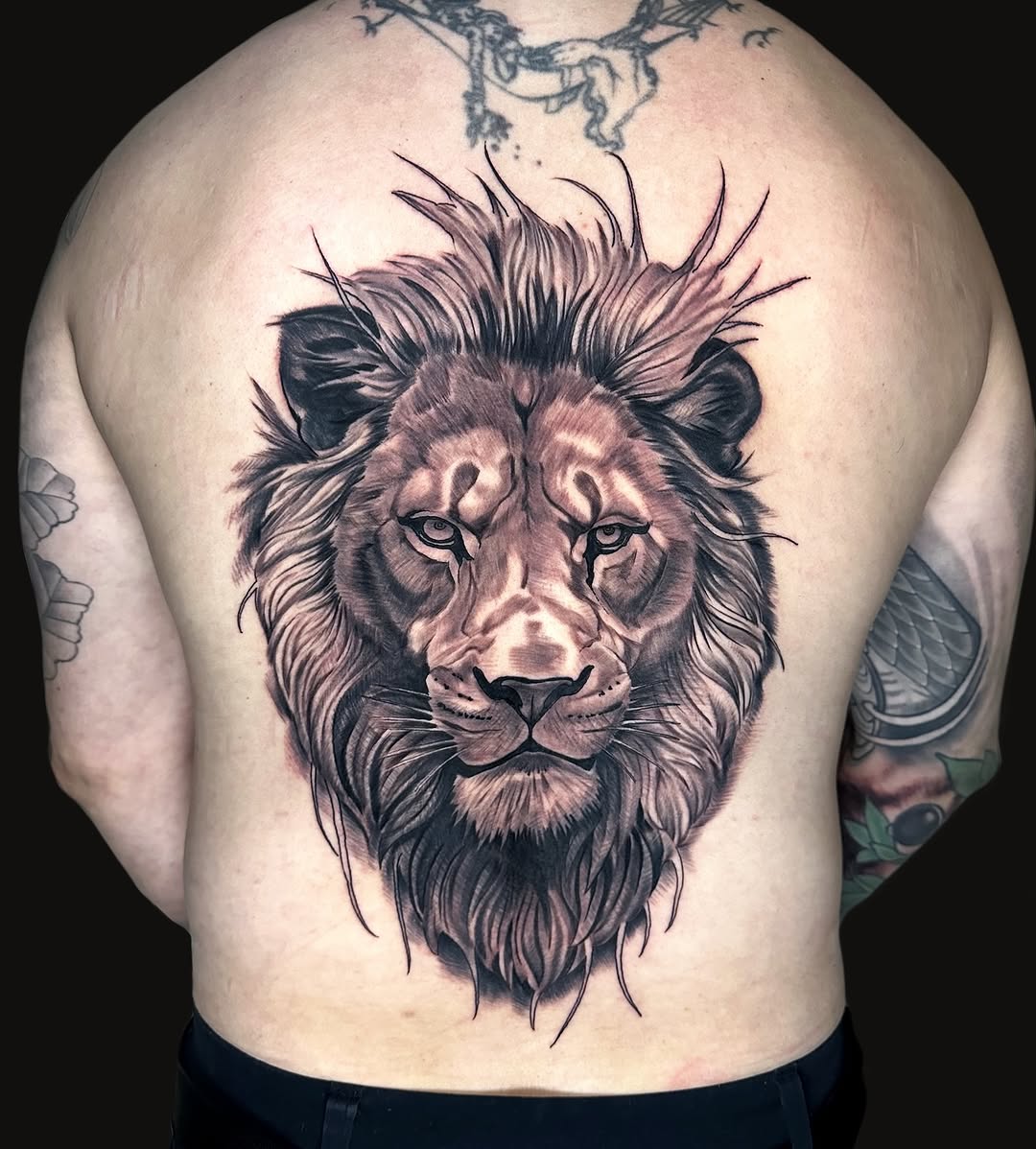 Lion head with flowing mane and detailed facial features in purple and black ink on the upper back of a shirtless man.