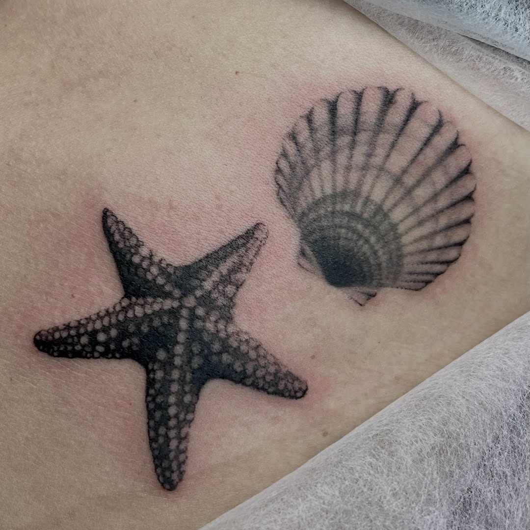 Starfish and scallop shell tattoo in black ink on pale skin, positioned on the upper arm or shoulder area.