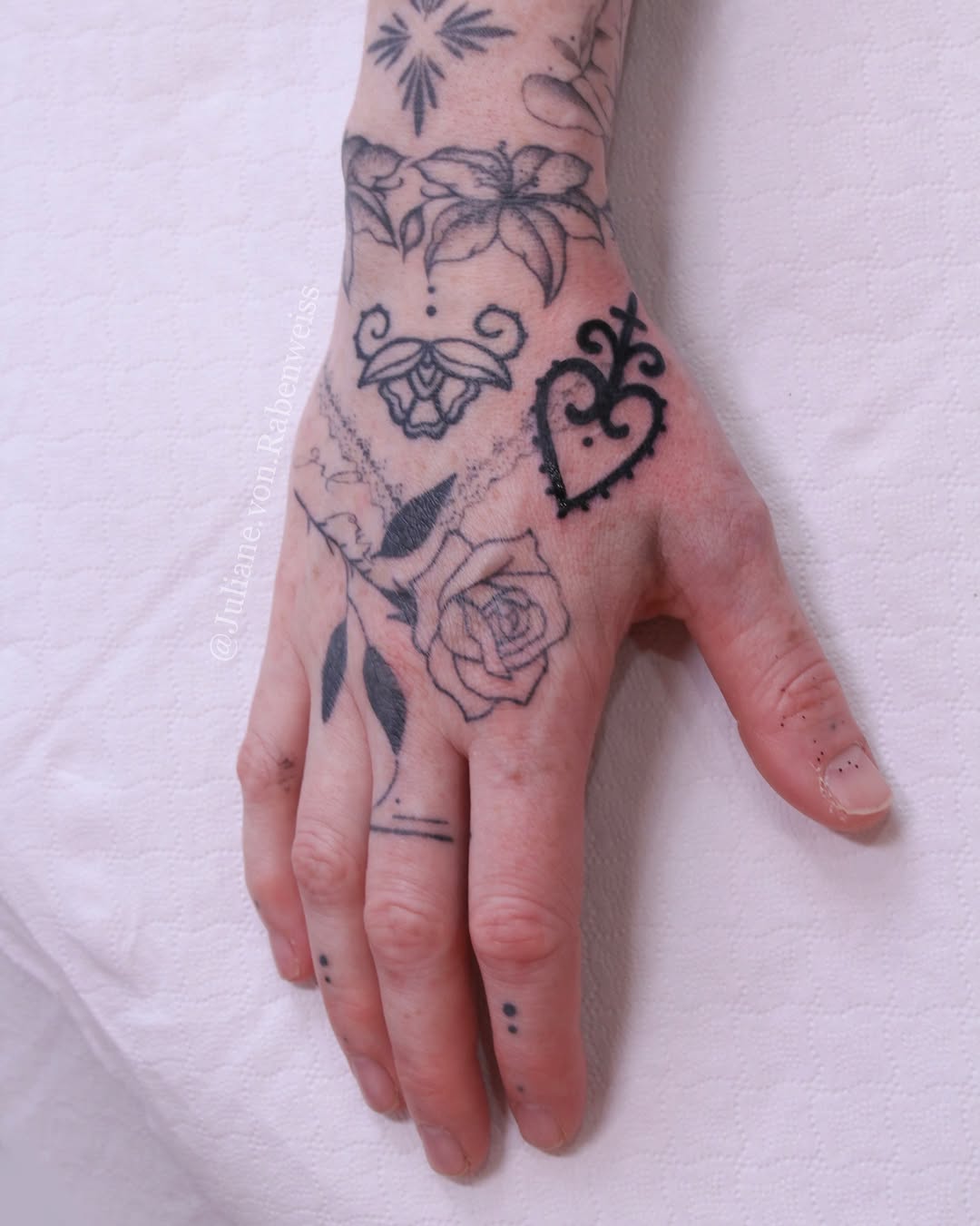 Hand tattoo featuring a decorative heart, rose, butterfly, flower motifs, and geometric designs in black ink on pale skin.