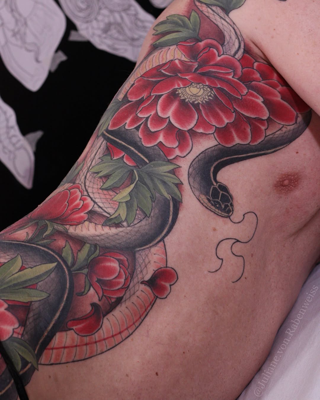 Colorful floral tattoo on the shoulder and chest featuring red flowers with green leaves and a black serpent, displayed on light skin.