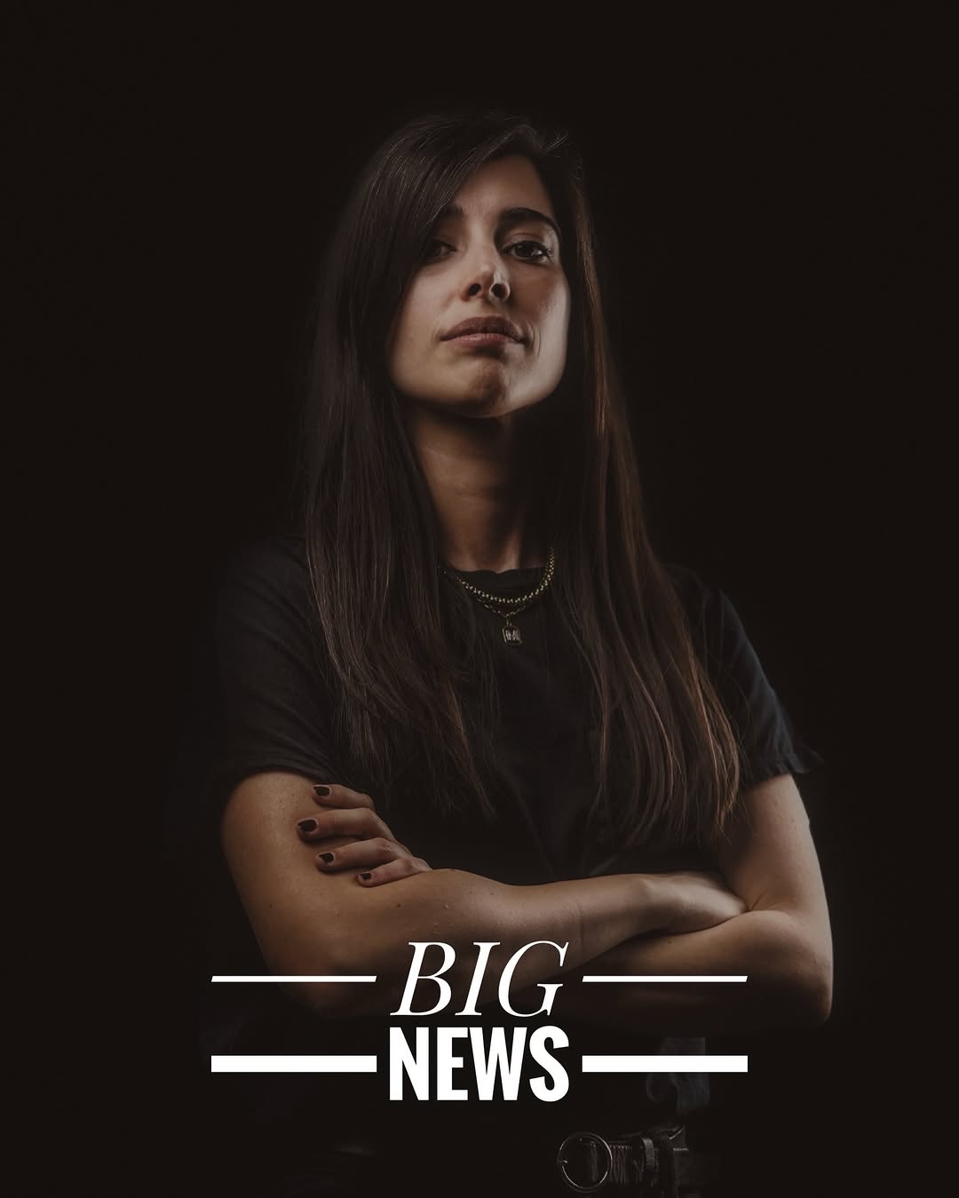Portrait photograph of a woman with long dark hair wearing a black shirt, displaying a small tattoo on her wrist against a dark background.