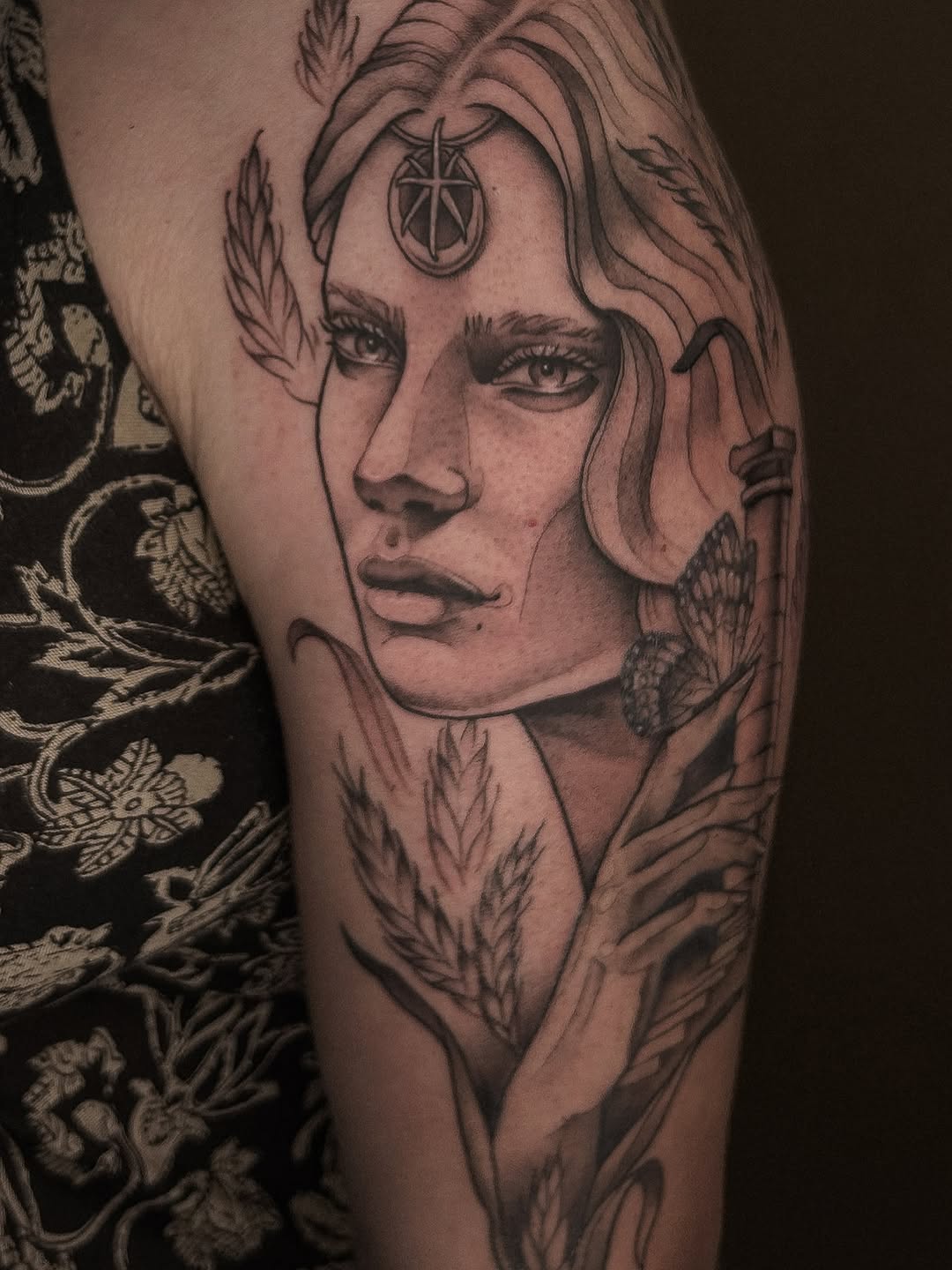 Portrait tattoo on upper arm featuring a face with closed eyes, ornamental third eye symbol on forehead, surrounded by feathered and botanical elements in black ink.