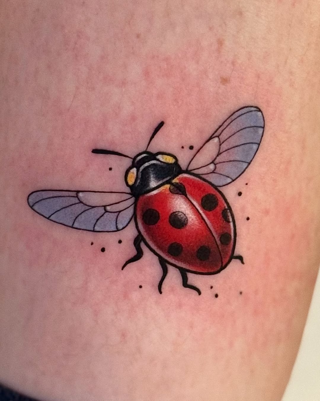 Red ladybug with black spots and translucent wings on human skin, featuring yellow head details and small decorative dots.