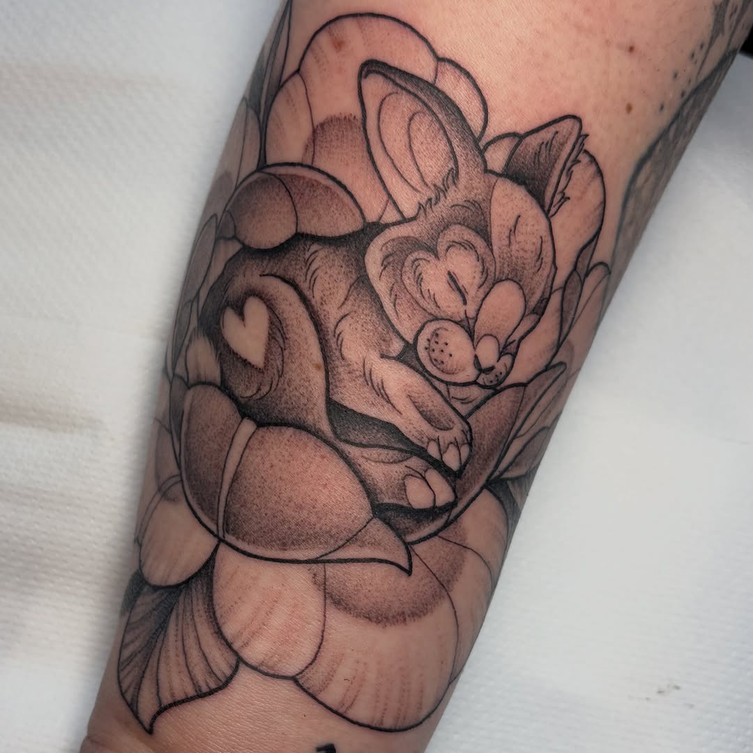 Floral arrangement tattoo featuring a smiling mouse or rat surrounded by layered petals and botanical elements in brown and black ink on the forearm.