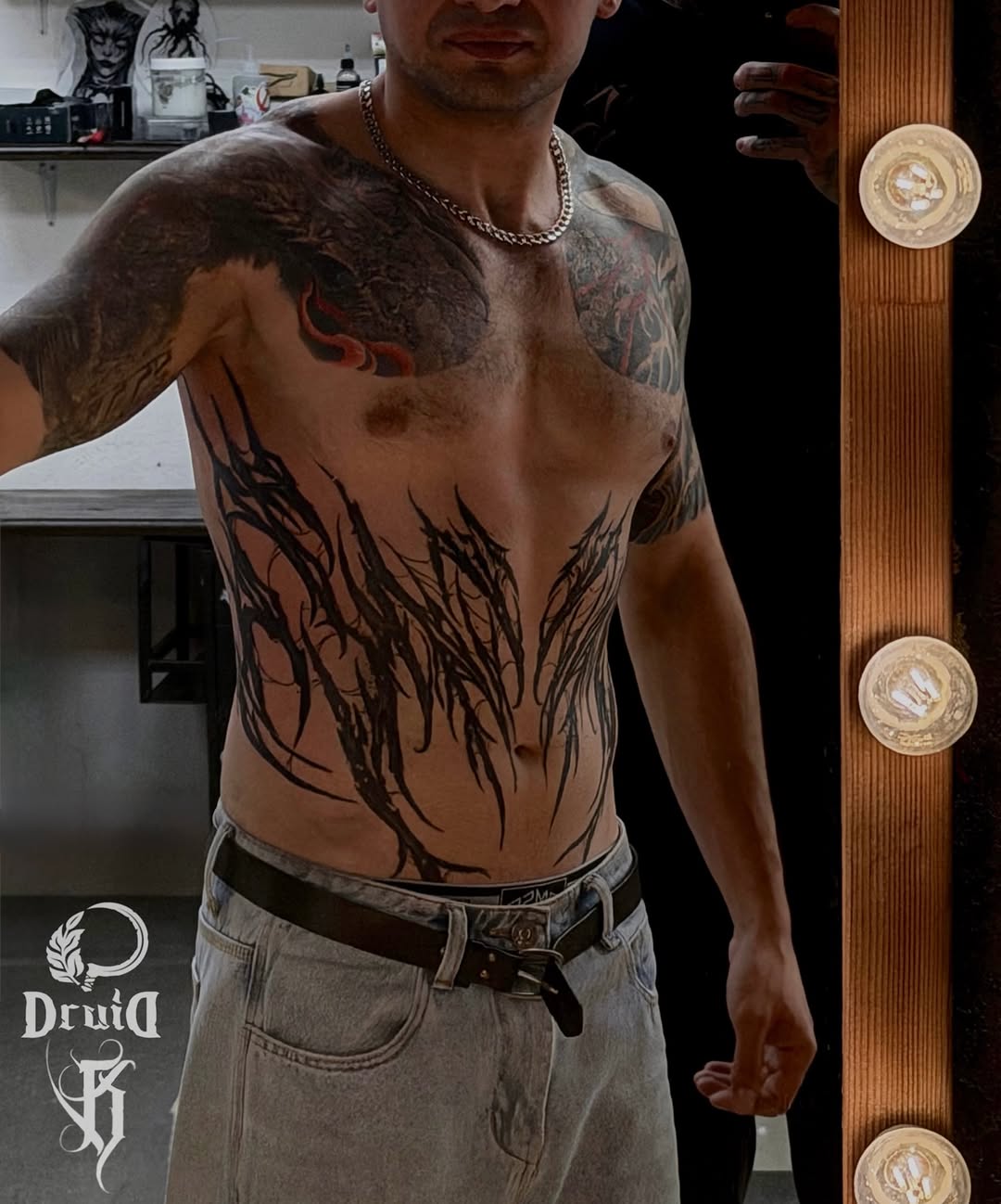 Shirtless man displaying large black and red chest and torso tattoo featuring abstract flowing lines and imagery, wearing jeans and chain necklace.