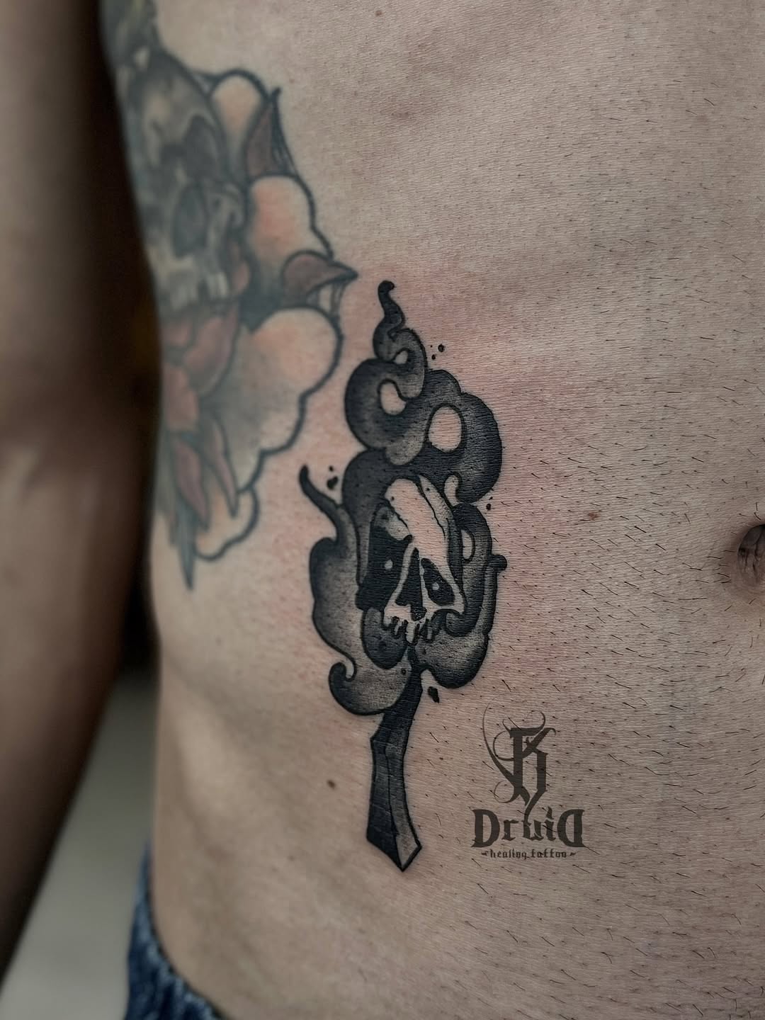 Black ink tattoo on the ribs featuring intertwined snakes with a skull, accompanied by a dagger and artist's logo and text below.