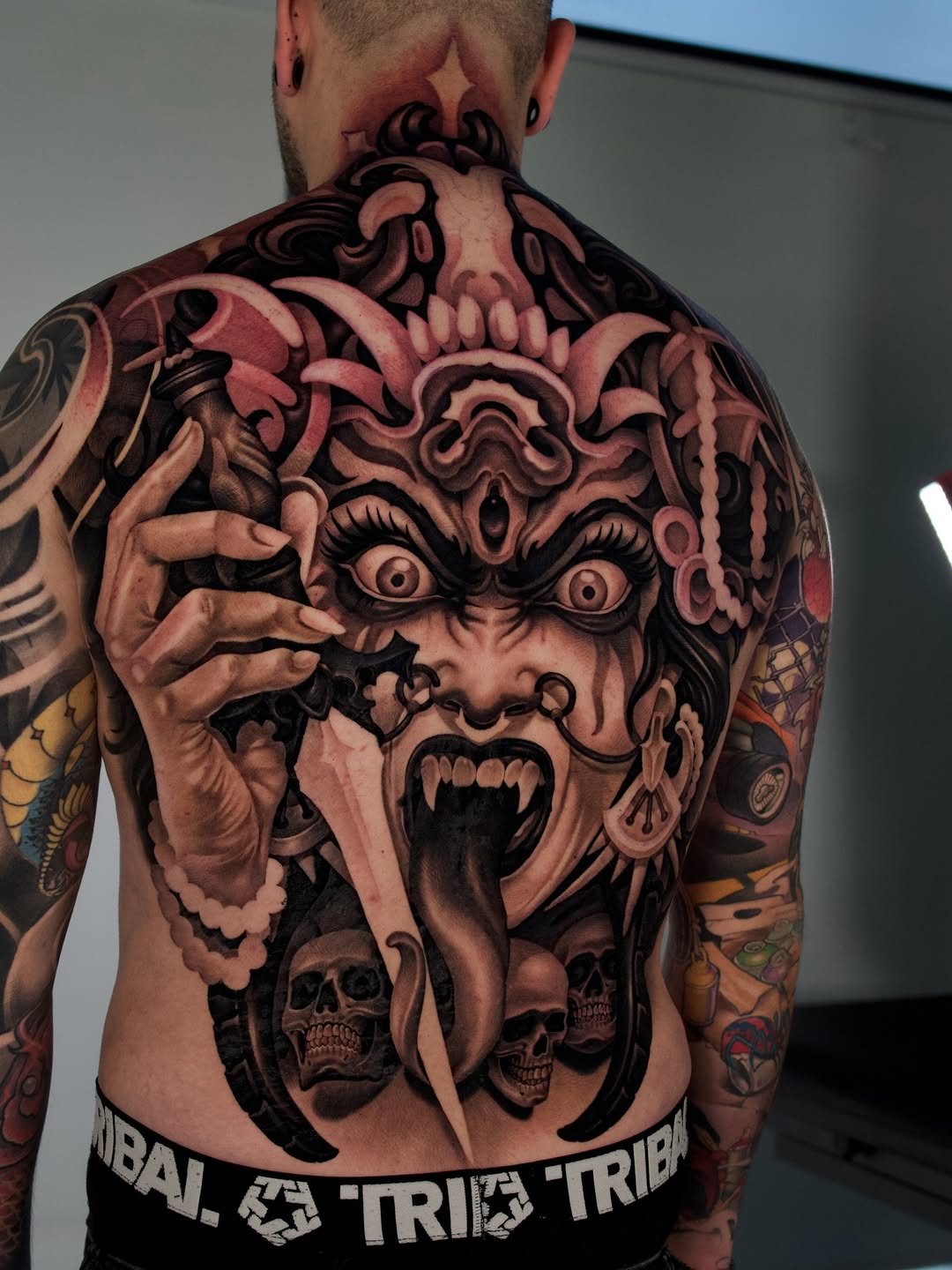 Back tattoo featuring a demonic face with elaborate horns, wide eyes, and open mouth, with skulls and ornamental details below, rendered in red and black ink.