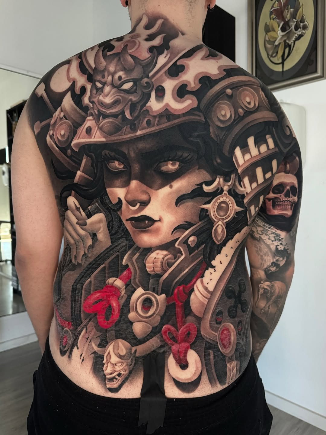 Back piece tattoo featuring a masked female face with ornate headdress, demons, skulls, and red heart designs in brown and red tones.