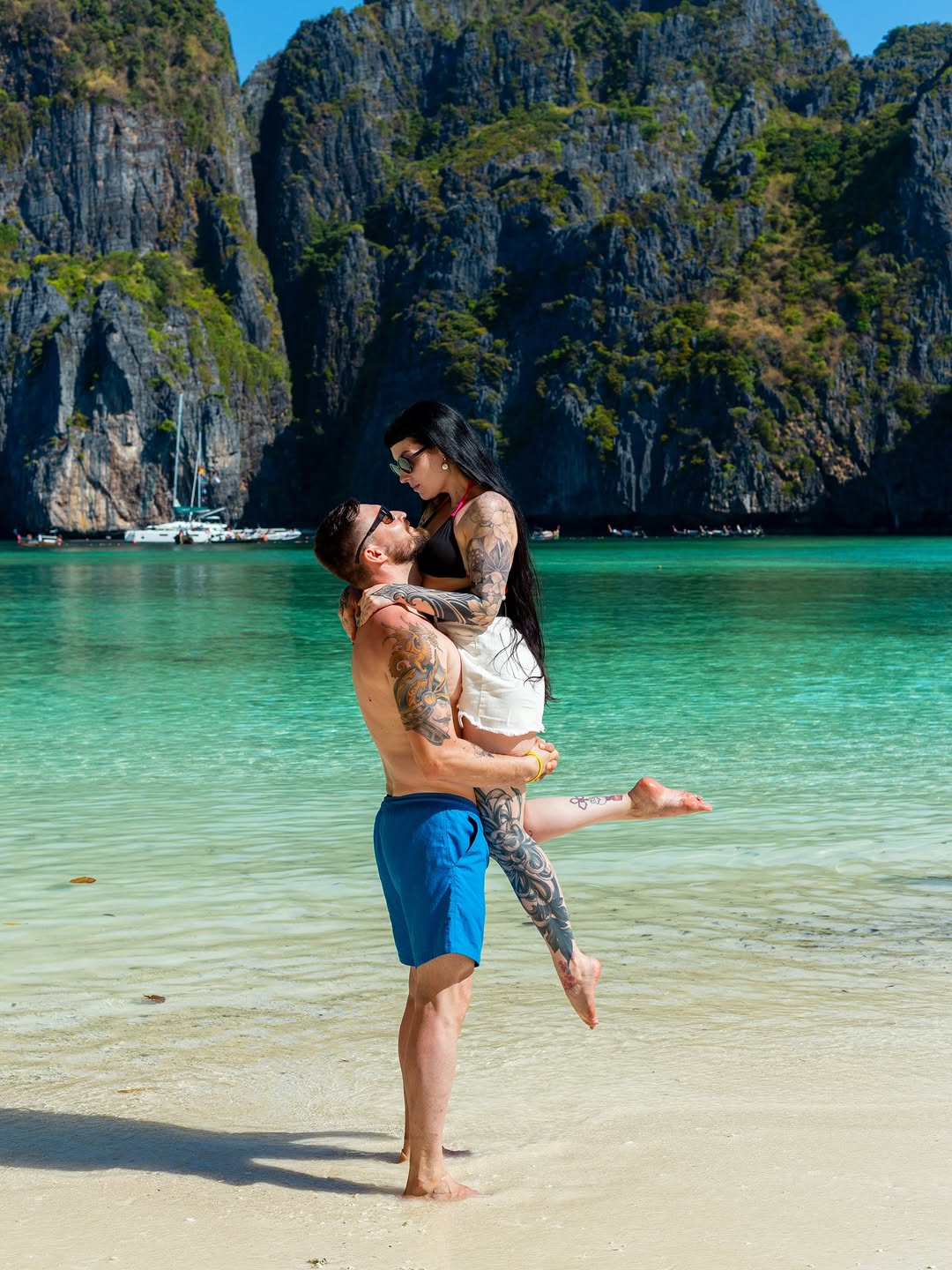 Tattooed man and woman embracing in turquoise tropical water with limestone cliffs in background, displaying visible arm and shoulder tattoos in black ink.