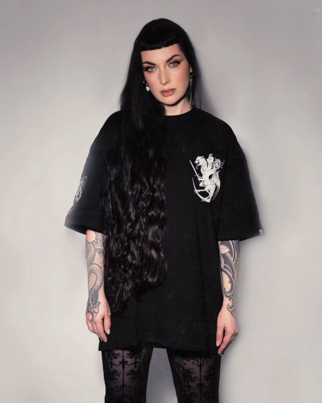 Ornate scrollwork tattoos visible on both forearms of a woman with long black hair wearing a black graphic t-shirt.