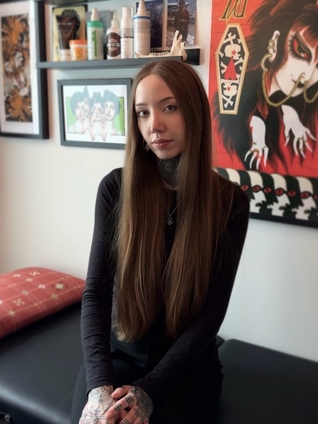 Portrait of a tattoo artist with long brown hair wearing black, displaying hand and finger tattoos in a studio with artwork on walls and shelves.