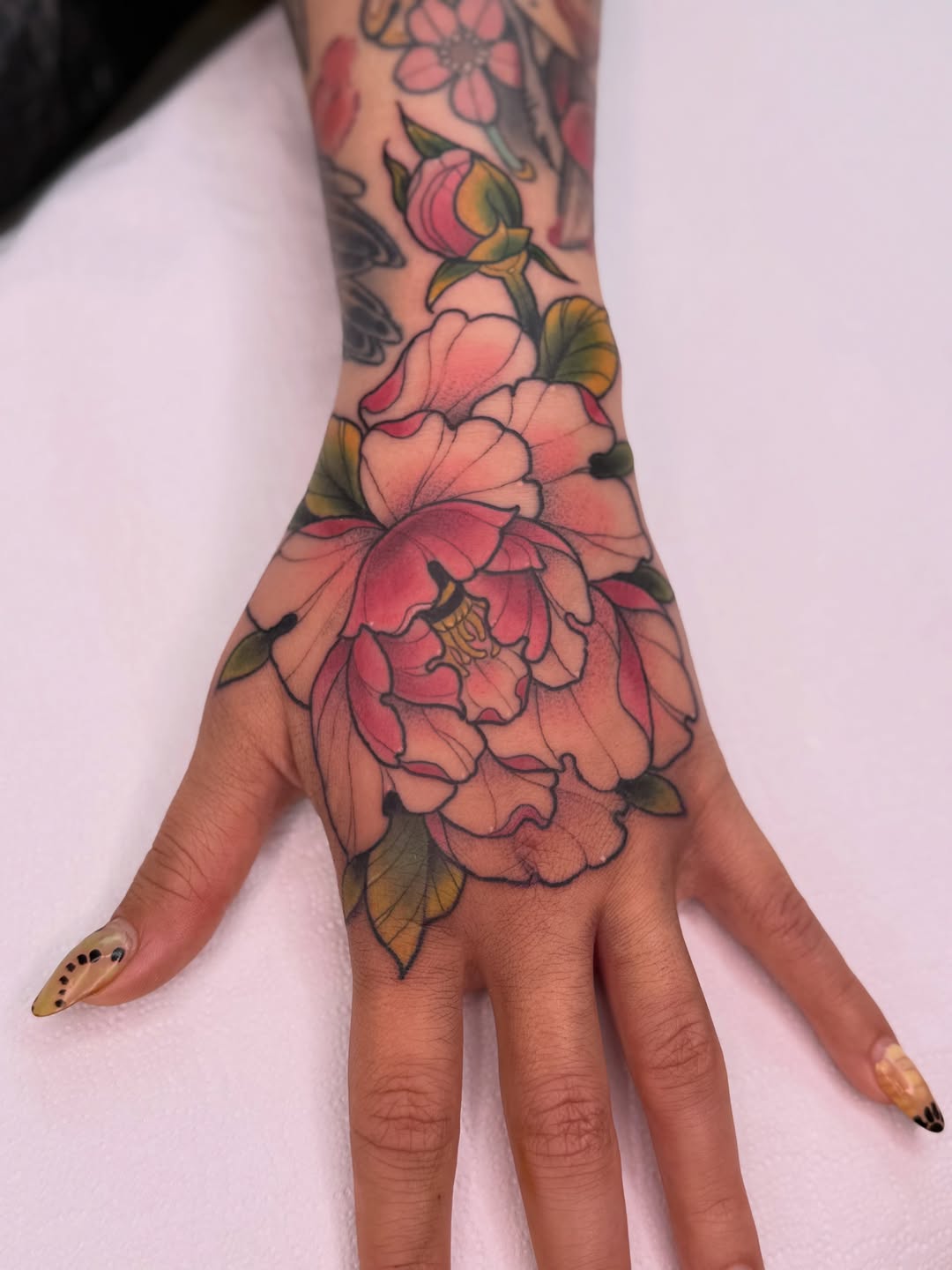 Colorful floral tattoo on hand and forearm featuring pink and coral blooms with green leaves and yellow accents, extending from wrist to fingers.