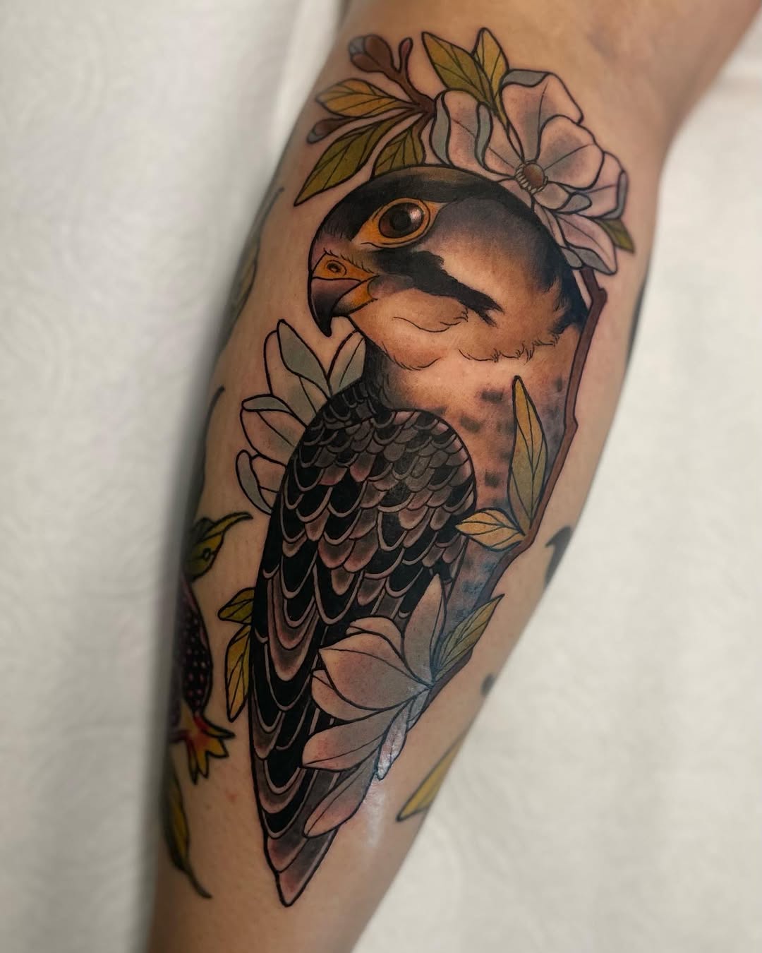 Falcon tattoo on the calf with detailed feather work, surrounded by pink and white flowers with green leaves in warm earth tones.