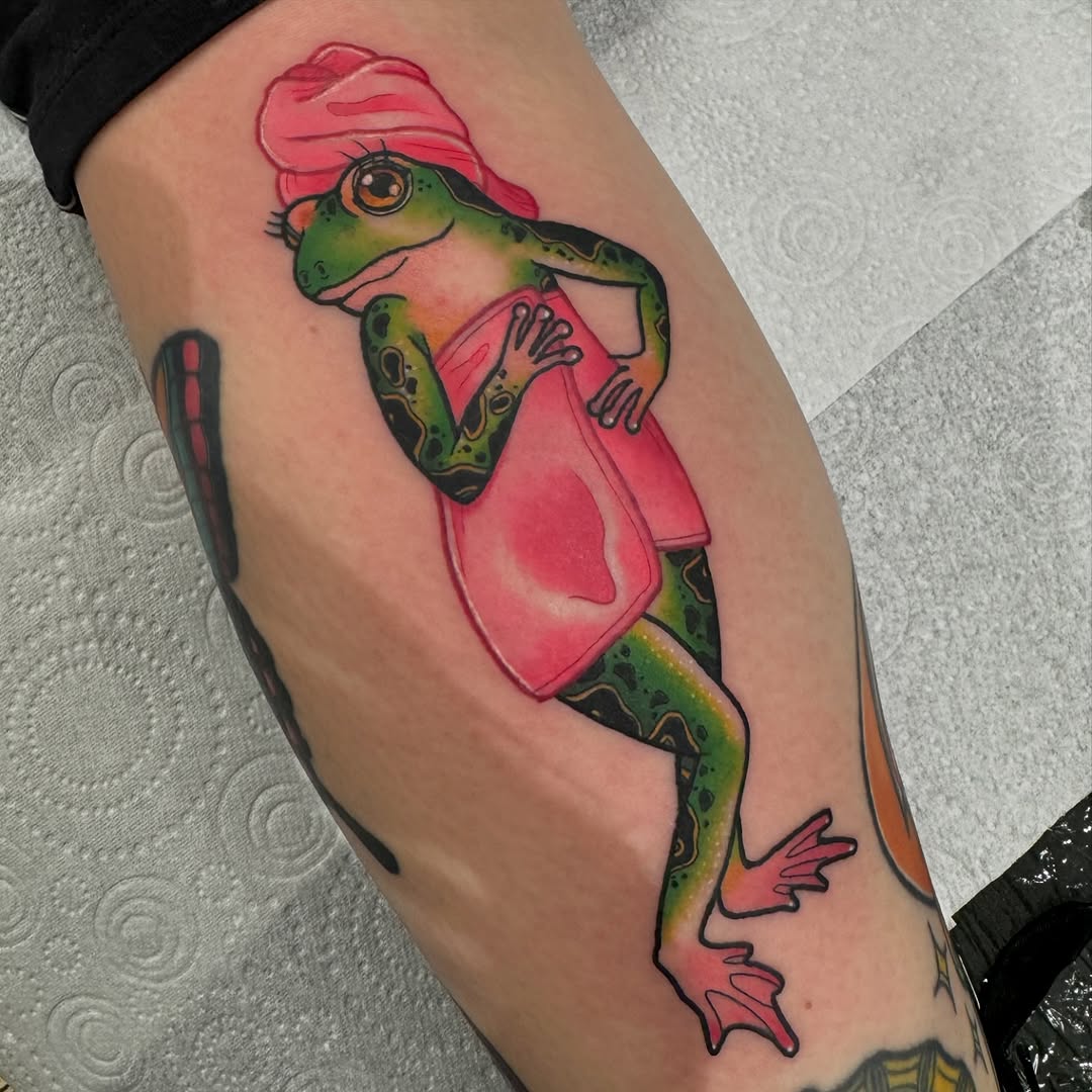 Colorful frog wearing a pink cap and jacket tattooed on the upper leg, featuring green, orange, and pink hues.