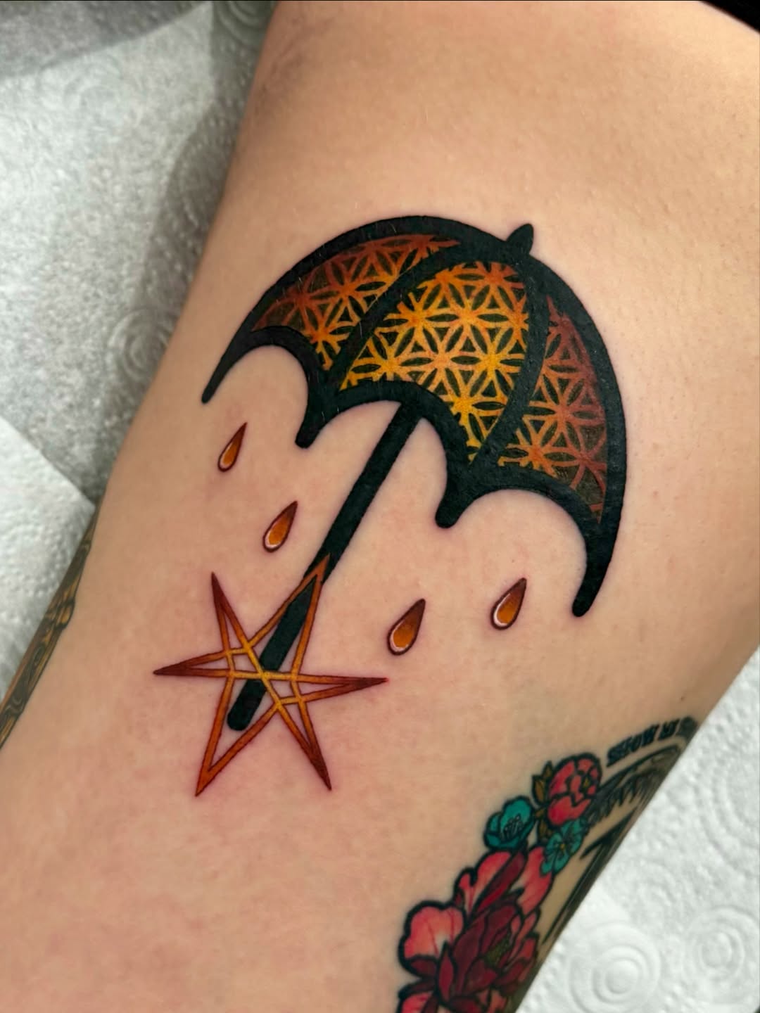 Umbrella tattoo with geometric patterned canopy in yellow and orange, falling raindrops, and a star below, on the upper arm.