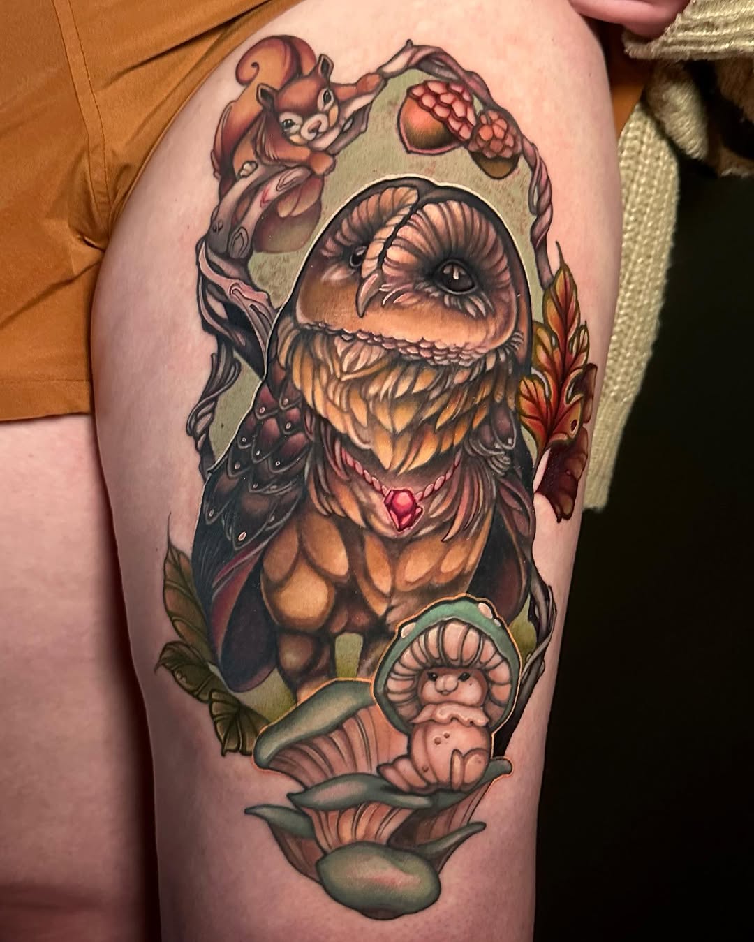 Colorful thigh tattoo featuring a large bearded creature with a heart on its chest, surrounded by smaller characters, mushrooms, and foliage in orange, brown, and green tones.