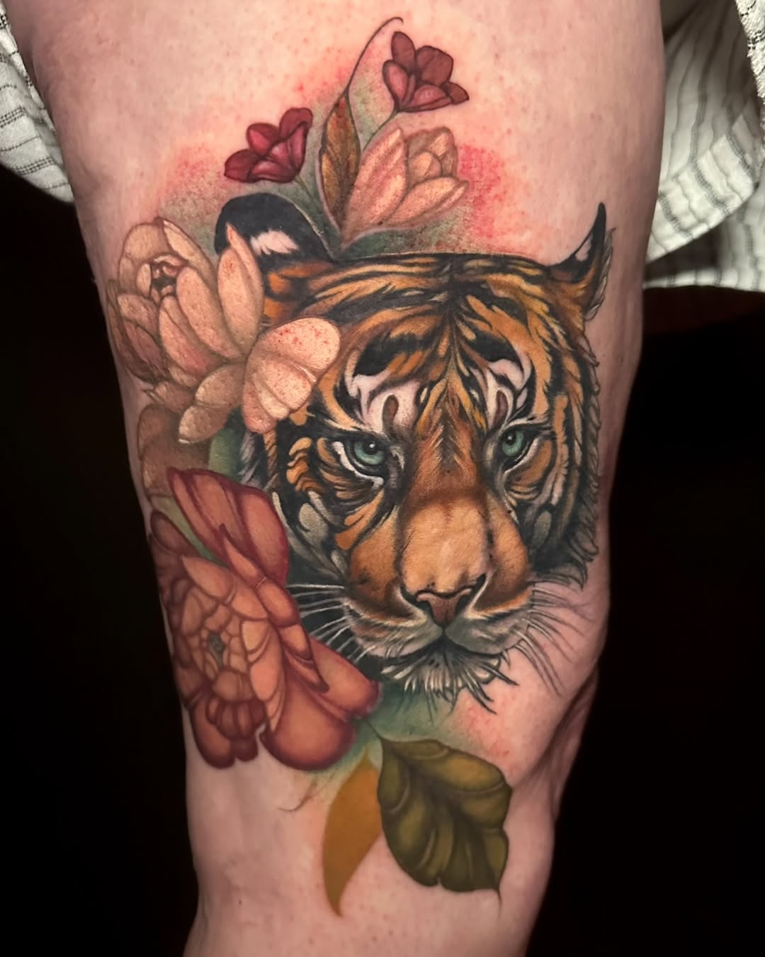 Tiger head with green eyes surrounded by pink and red flowers on the thigh, featuring orange and black stripes with green foliage.