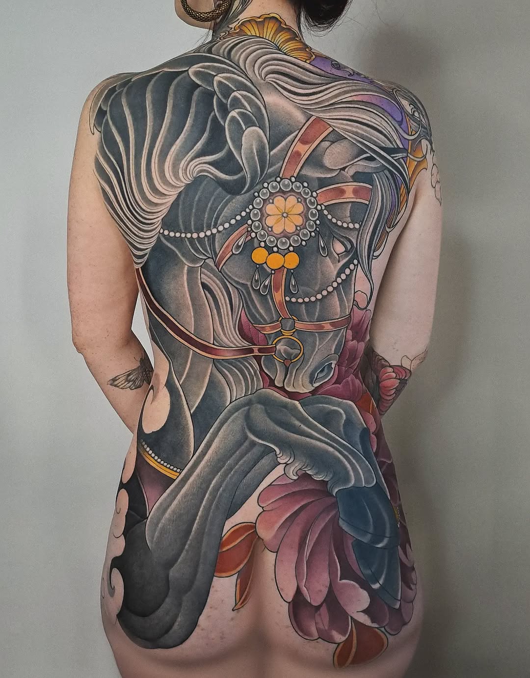 Back tattoo featuring an ornate horse head with flowing mane, decorative bridle, and floral elements in purple, gold, orange, and burgundy tones.