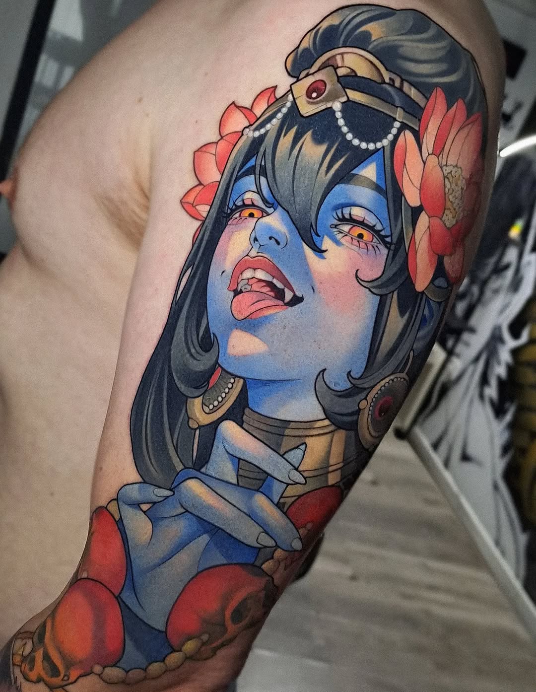 Blue-skinned female figure with dark hair, gold headpiece, red flowers, and gold jewelry tattooed on upper arm in vibrant colors.