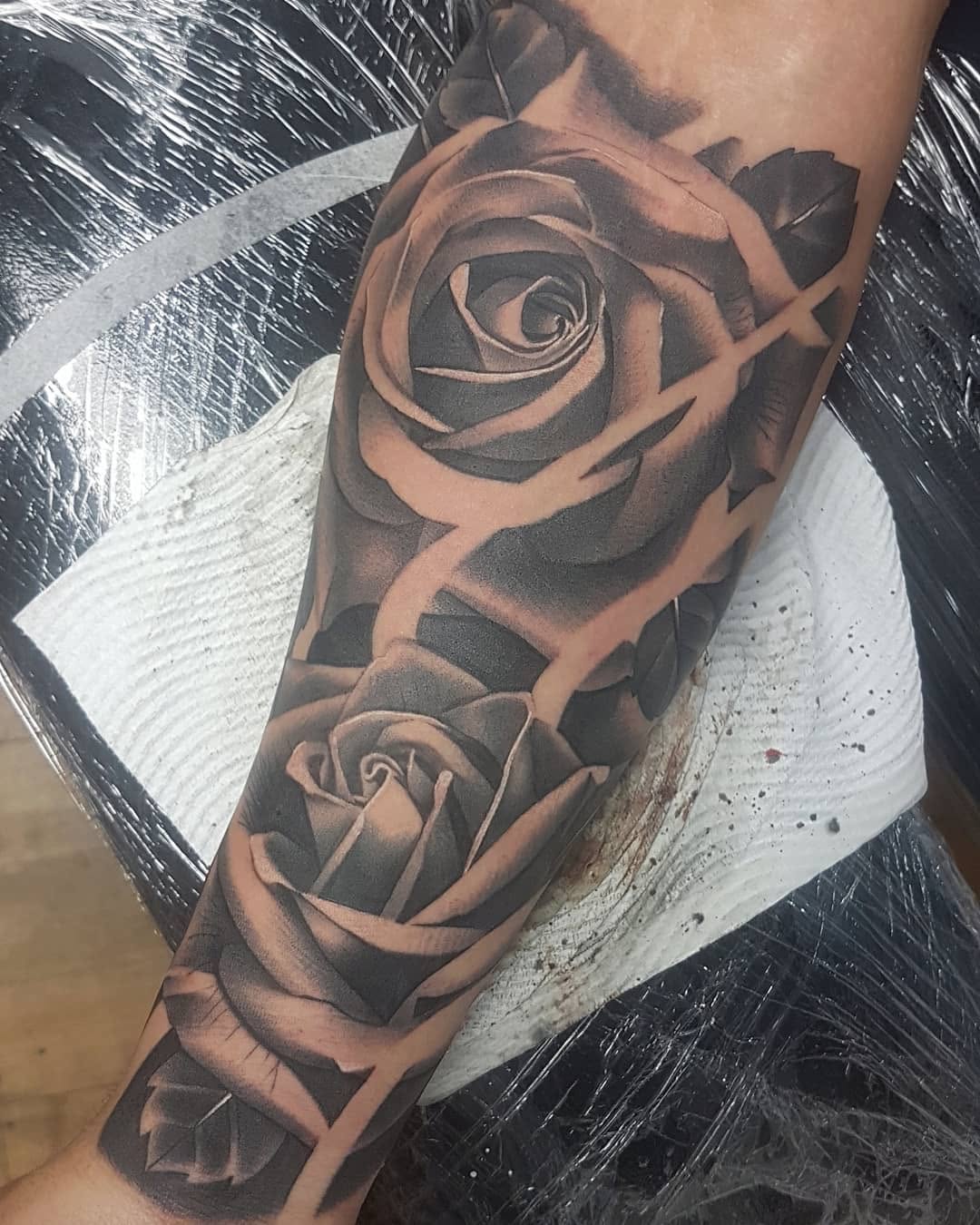 Three roses in varying sizes with detailed shading on the forearm, rendered in grayscale tones on skin with a white bandage visible.