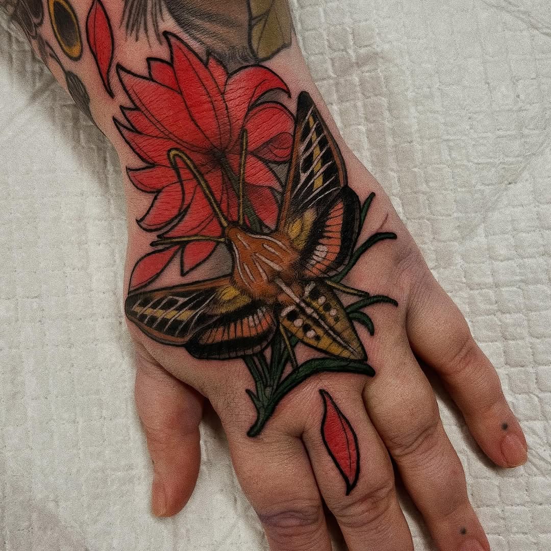 Colorful butterfly with brown and gold wings resting on a bright red flower, tattooed on the back of a hand with additional red petals on the fingers.